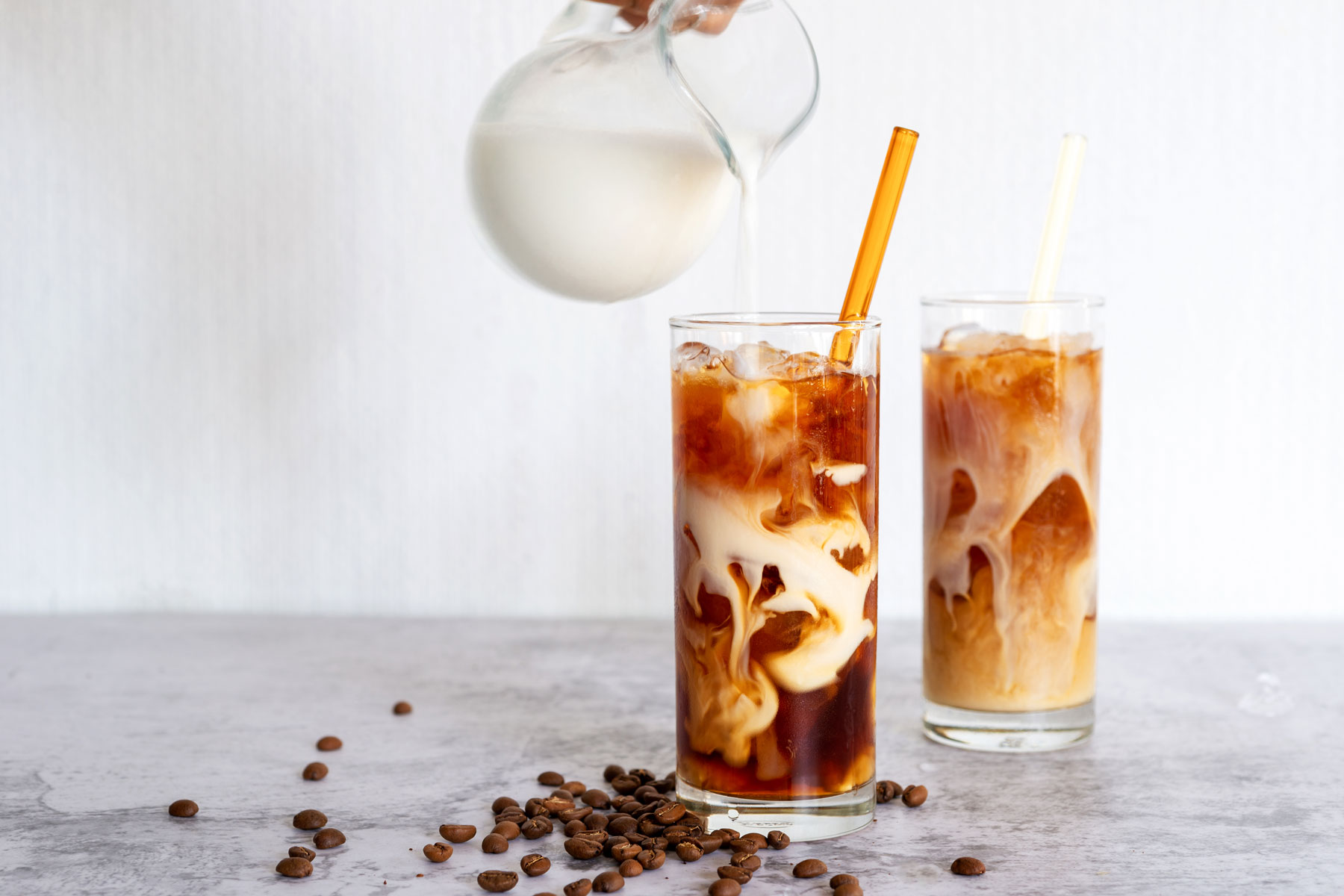 A pitcher of milk being poured into two glasses of iced coffee